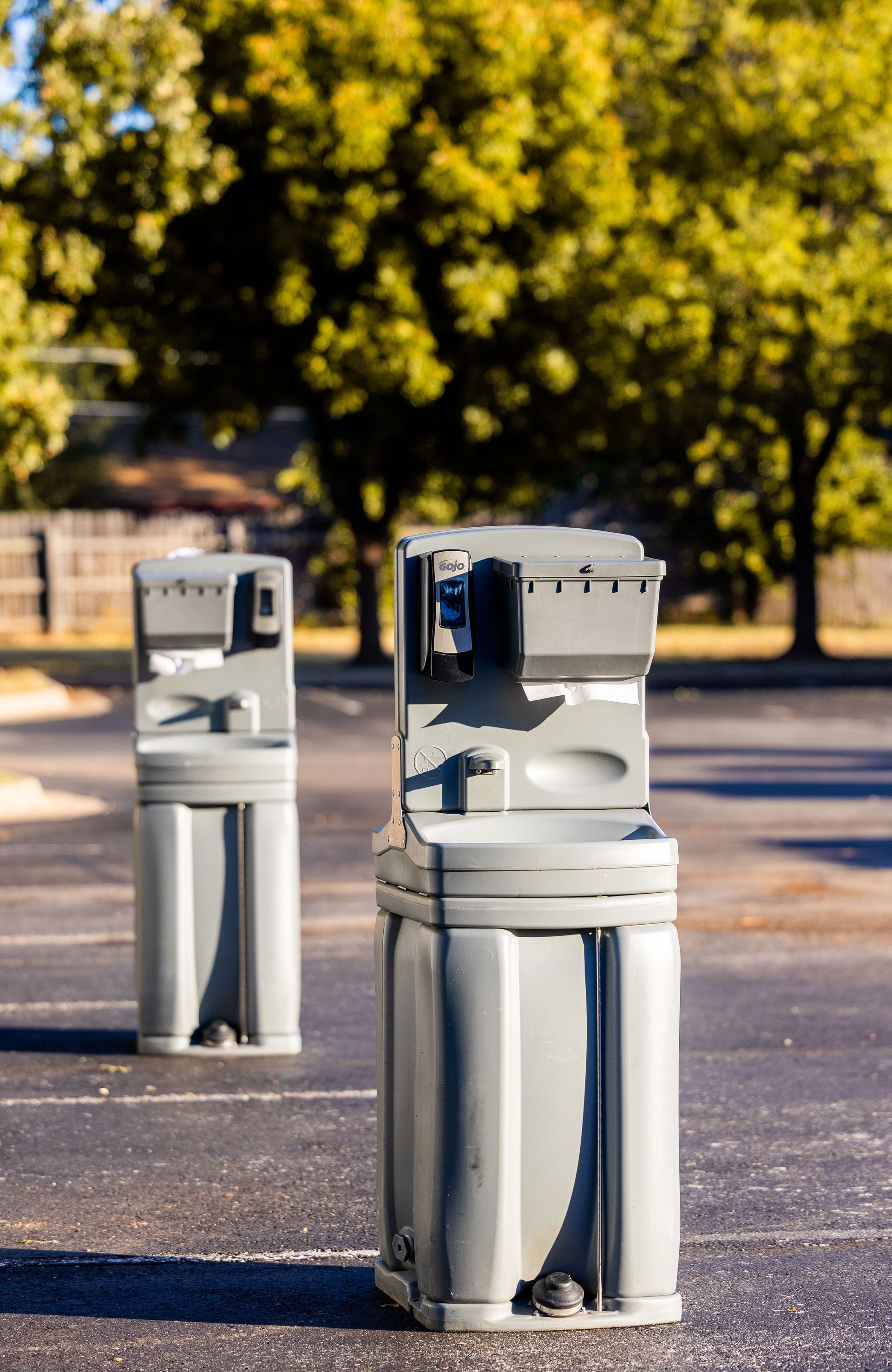 Outdoor portable hand washing stations on asphalt with trees in the background.